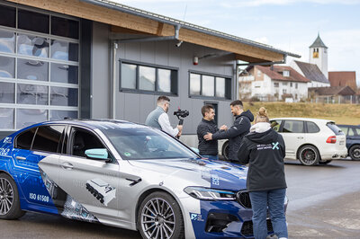 Mehrere Menschen stehen um einen gebrandetes Auto von Arnold NextG. Zwei Männer geben sich die Hand, ein weiterer Mann hält mit einer Kamera drauf und eine Junge Dame steht mit dem rücken zur Kamera.