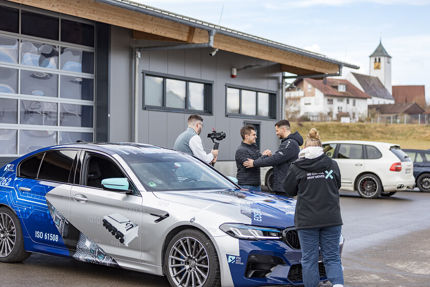 Mehrere Menschen stehen um einen gebrandetes Auto von Arnold NextG. Zwei Männer geben sich die Hand, ein weiterer Mann hält mit einer Kamera drauf und eine Junge Dame steht mit dem rücken zur Kamera.
