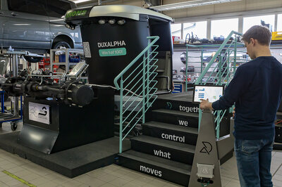Ein junger Mann steht am rechten Bildrand vor einer Stehle und drückt dort etwas an einem Touch Display. Im Hintergrund bzw. in der Mitte des Bildes sieht man eine dunkle kleine Treppe mit türkisenem Geländer und einer folierten schwarz-silbernen Traktor Kabine, davor befindet sich eine Traktor Vorderachse. 
