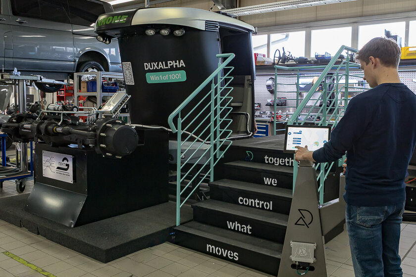 Ein junger Mann steht am rechten Bildrand vor einer Stehle und drückt dort etwas an einem Touch Display. Im Hintergrund bzw. in der Mitte des Bildes sieht man eine dunkle kleine Treppe mit türkisenem Geländer und einer folierten schwarz-silbernen Traktor Kabine, davor befindet sich eine Traktor Vorderachse. 