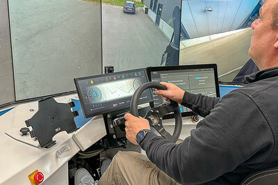 Ein Mann sitzt am rechten Bildrand vor mehreren großen Bildschirmen, die links zu sehen sind, auf denen eine Straße abgebildet ist. Vor dem Mann befindet sich ein Lenkrad mit zwei Touch Displays.