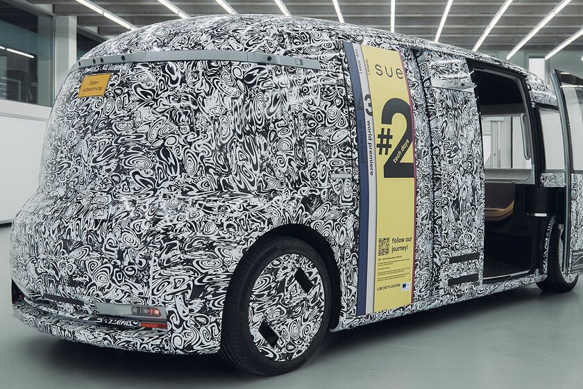 Ein noch getarnter Bus (schwarz-weißes Muster) steht in einer lehren beleuchteten hellen Halle. Der Bus hat keine Fenster.