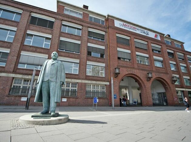 Standort Rüsselsheim. Eingang der Motorworld Rüsselsheim. Rotes Backstein Gebäude mit einer Statue davor.