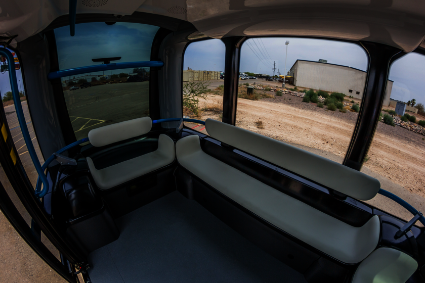 Ein Innenraum eines Fahrzeuges, welches rundherum Fenster besitzt, mit weißen Bänken davor. Dieses Fahrzeug hat kein Lenkrad. In der Mitte befindet sich Platz zum stehen.  