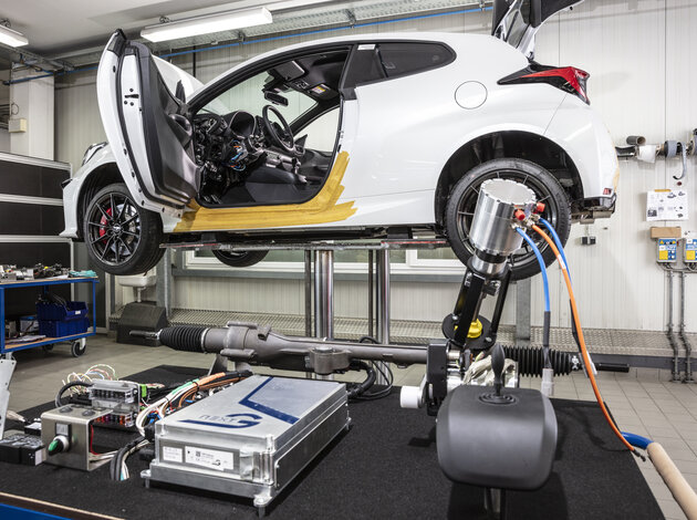 Ein auto ist auf einer Hebebühne in einer Werkstattt hochgefahren, die Fahrertürsteht offen und man sieht das am Lenkrad was neu verbaut ist. Im Vordergrund steht ein Tisch auf dem alle Komponenten von NX NextMotion liegen (Steuergerät, Joystick, Lenksäule mit Motor).