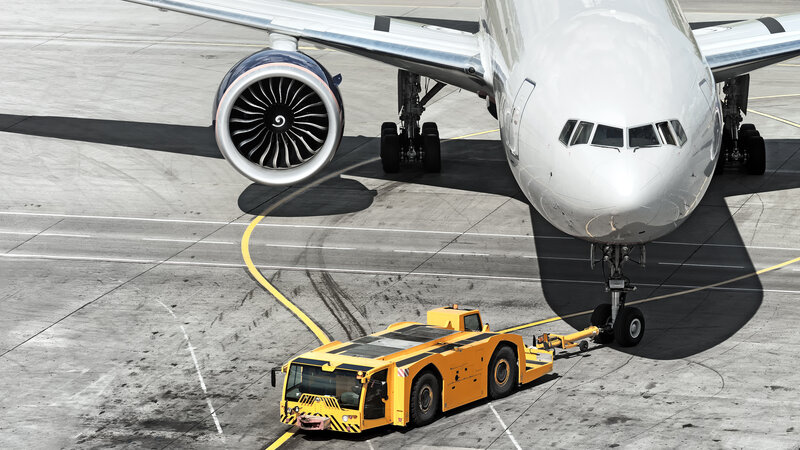 Auf einem Flughafen Rollfeld zieht ein sogennater Pusher, also ein gelber Flugzeugschlepper, ein großes Flugzeug von rechts oben nach links vorne.
