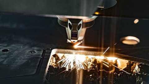 Eine Machine lasert in eine dicke Metallplatte linien rein. Unter der Platte sieht man kleine Orange/Gelbe funken.