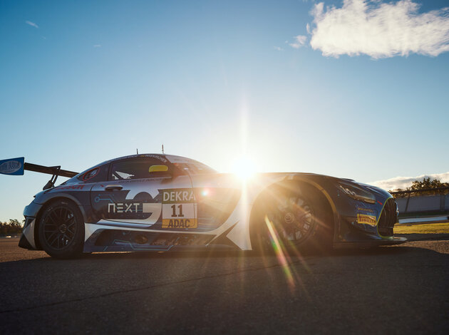 Ein Mercedes GT3 steht von links nach rechts, bei blauem Himmel und Sonnenschein auf einem geterten Platz. Die Sonne ist zur hälfte über der Motorhaube zu sehen.