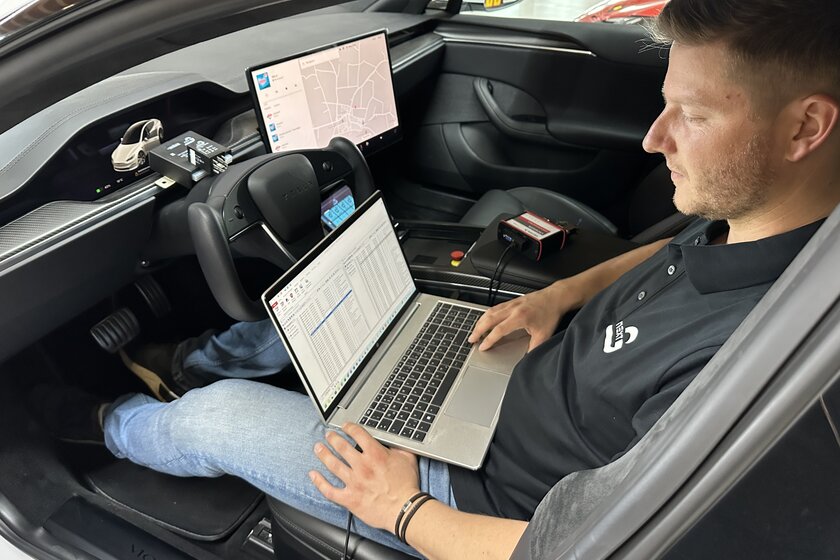 Ein Mann sitzt in einem Auto und hat vor sich seinen Laptop auf dem ein Softwareprogramm läuft. Neben ihm und vor ihm liegen noch verschiedene Gateways und Kabel.