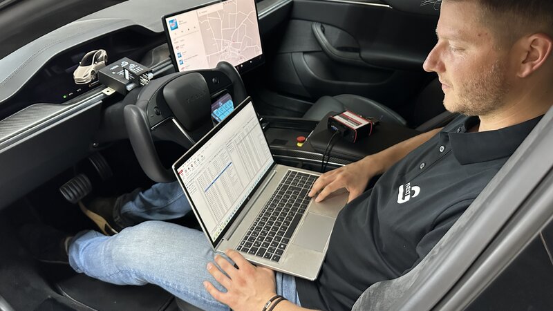 Ein Mann sitzt in einem Auto und hat vor sich seinen Laptop auf dem ein Softwareprogramm läuft. Neben ihm und vor ihm liegen noch verschiedene Gateways und Kabel.