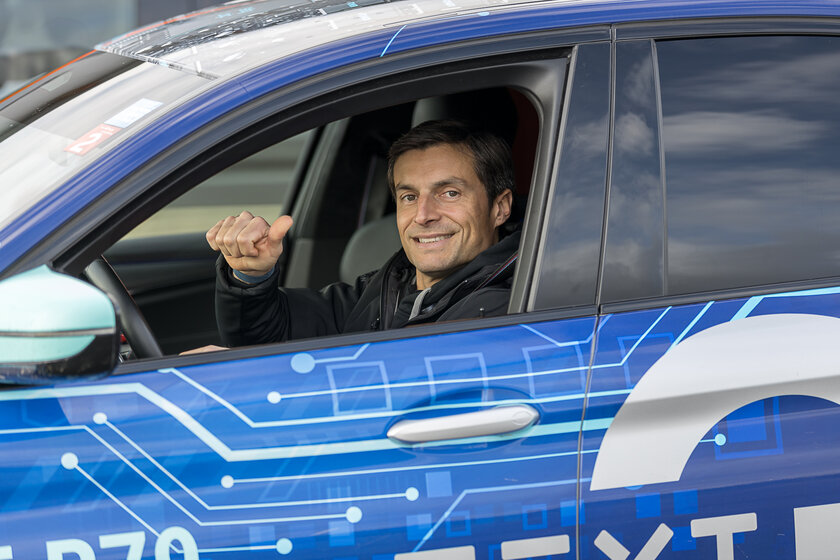 In einem gebrandeten blauen Auto ist bei der Fahrertür das Fenster unten. Im Auto sitzt ein lächelnder braunhaariger Mann und schaut aus dem Fenster. 