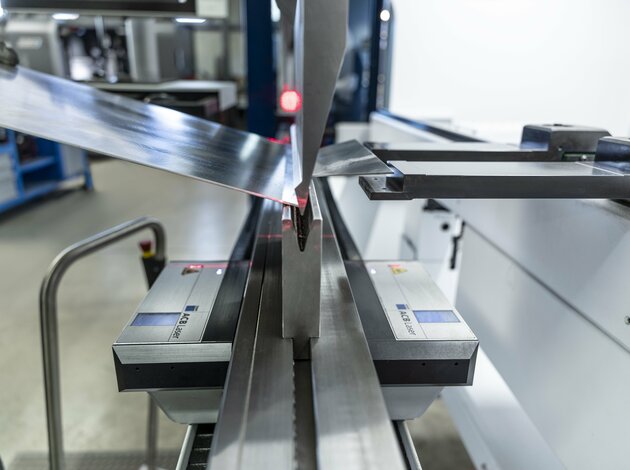 In einer Maschine wir eine Metallplatte gebogen. Im Hintergrund erkennt man unscharf noch ein rotes Licht.