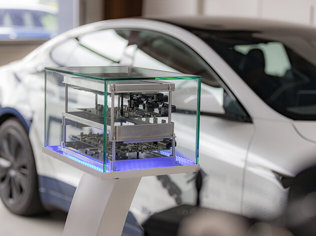Im Vordergrund befindet sich das NX NextMotion, welches auseinandergezogen auf 3 Ebenen aufgebaut ist, sodass die Platinen im inneren zum vorschein kommen. Das ganze wird in einer Glasvitrine präsentiert. Im Hintergrund steht unscharf ein weißer Tesla.