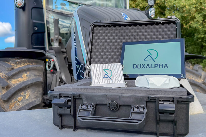 Ein Traktor steht unscharf im Hintergrund. Im Vordergrund steht ein geöffneter schwarzer Koffer in dem ein Touch Display steht mit DuxAlpha Logo drauf, links daneben liegt eine DuxAlpha gebrandete ECU und davor eine weiße Antenne.