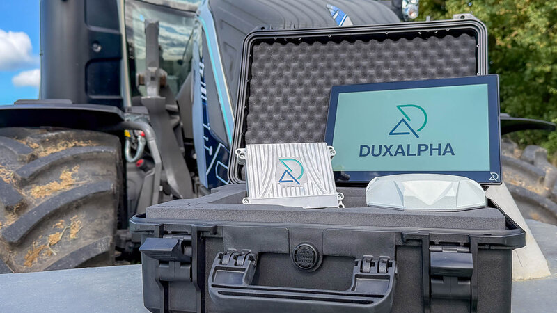 Ein Traktor steht unscharf im Hintergrund. Im Vordergrund steht ein geöffneter schwarzer Koffer in dem ein Touch Display steht mit DuxAlpha Logo drauf, links daneben liegt eine DuxAlpha gebrandete ECU und davor eine weiße Antenne.