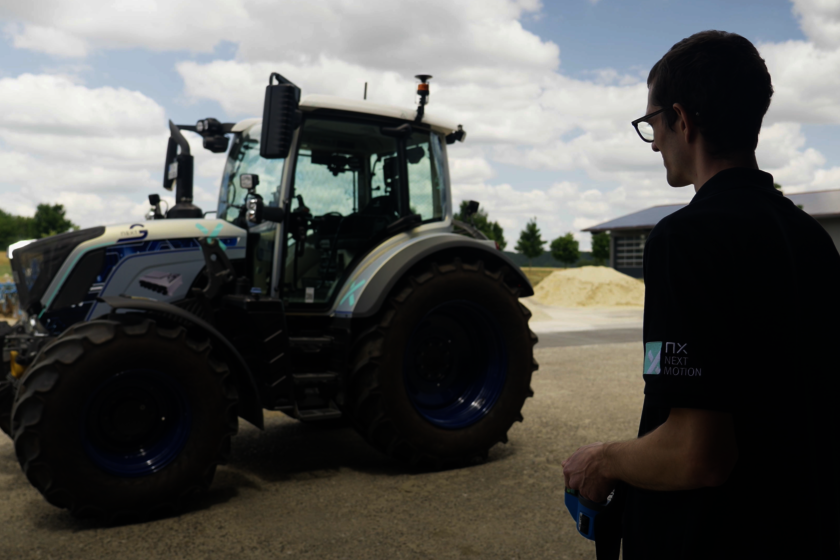 Ein Mann steht vor einem silbernen blauen Traktor, in dem keiner drin sitzt. Der Mann der am rechten Bildrand zu sehen ist, steht mit einer Fernsteuerung in der Hand und steuert den Traktor.  