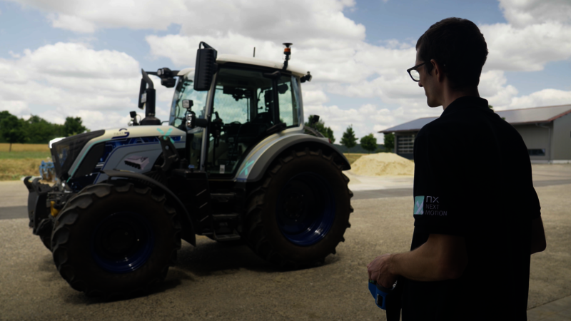 Ein Mann steht vor einem silbernen blauen Traktor, in dem keiner drin sitzt. Der Mann der am rechten Bildrand zu sehen ist, steht mit einer Fernsteuerung in der Hand und steuert den Traktor.  