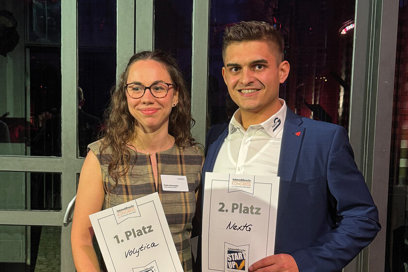 Zwei Menschen stehen mit einer Urkaunde lächelnd da. Links eine Dame in einem braunen Kleid, rechts ein junger attraktiver strahlender Mann in einem weiß blauen Anzug.