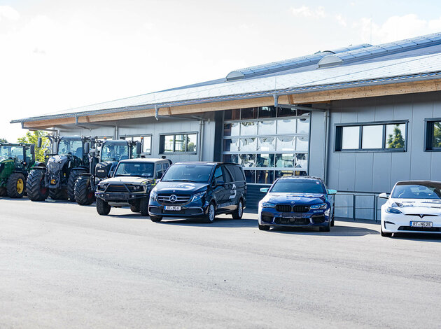 Traktoren, Geländewagen, Transporter und Pkw – darunter Mercedes, BMW und Tesla – geparkt vor einem modernen Firmengebäude mit Garagentor.