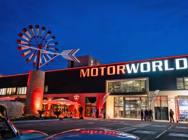 Standort Mallorca. Der Parkplatz und der Eingang der Motorworld Mallorca bei Nacht, bei dem sehr viel leuchtet rot und weiß leuchtet.