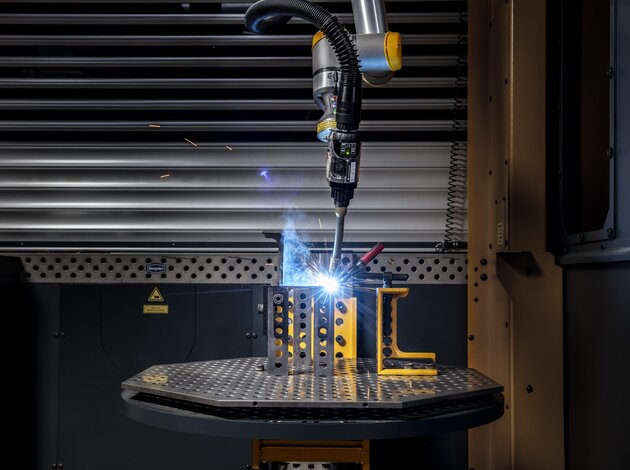 Ein Schweißroboter schweißt auf einer Platte zwei metallstücke zusammen. Beide liegen im 90 grad Winkel zueinander und in dieser Ecke schweist er. Aus dem Roboter Schweißarm erscheint blaues grelles Licht durchs schweißen.
