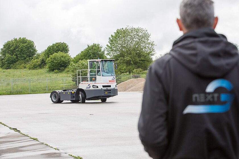 Ein ferngesteuerter Terminal-Traktor der Firma MAFI, umgebaut mit NX NextMotion, bewegt sich durch eine Fernsteuerung über einen Übungsplatz. Im Vordergrund rechts steht ein Mitarbeiter, mit Blick zum Terminal Traktor, der den MAFI steuert.