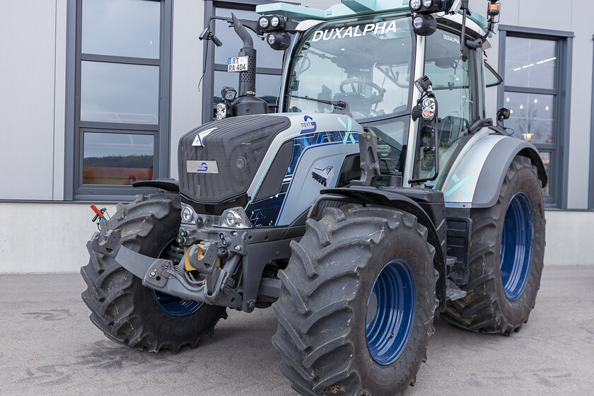Ein Silberner Blau Cyan-farbener Traktor steht vor einer Halle posierend. 