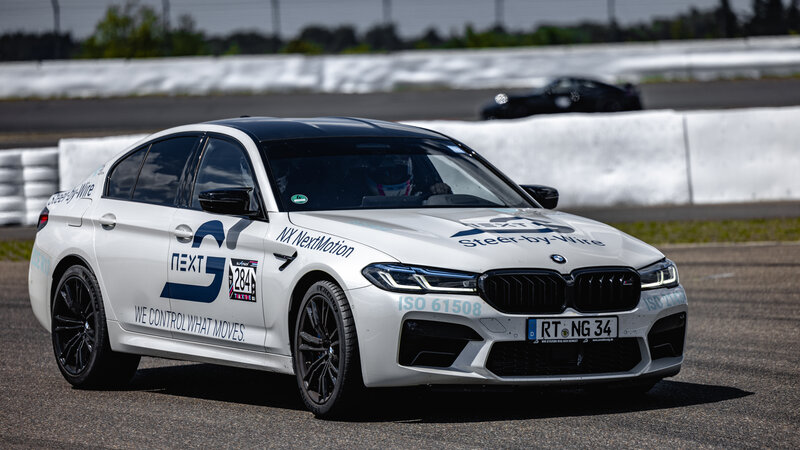 Ein weißer BMW M5 mit Arnold NextG Logo und Licht an fährt über eine Asphalt strecke.