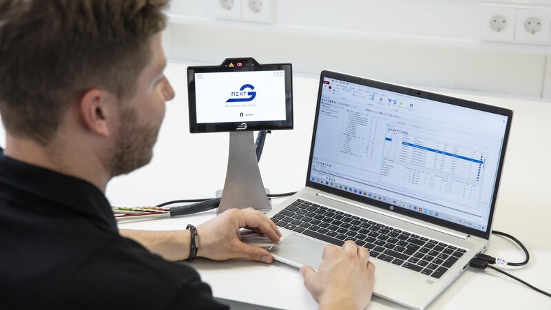 Ein blondbrauner Mann mit Bart sitzt vor seinem Laptop und arbeitet daran. Links neben dem Laptop steht ein Touch Display mit Arnold NextG Logo.