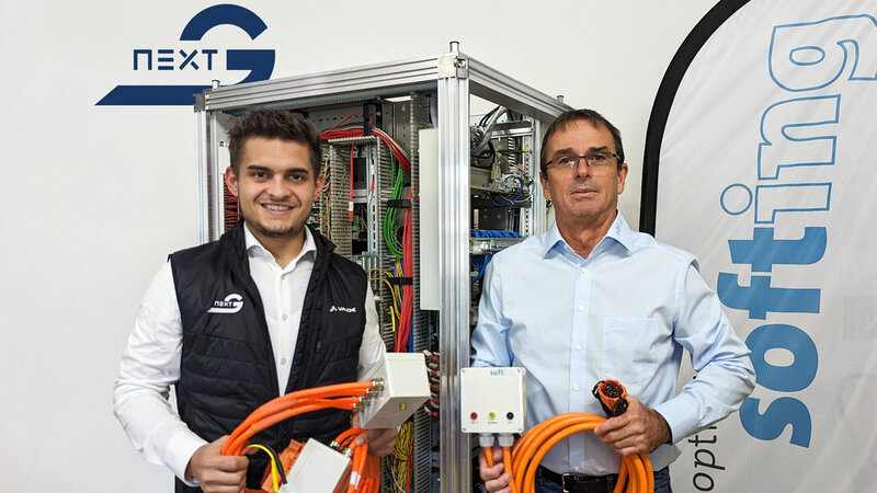 Zwei Männer stehen nebeneinander und jeder hat ein orangenes Kabel in der Hand. Im Hintergrund steht ein großer Technikschrank mit vielen Kabeln und eine Beachflag von softing.