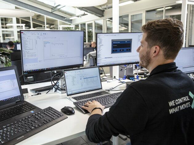 Ein Mann mit Bart und Arnold NextG Pulli sitzt an seinem Arbeitspplatz und arbeitet an seinen Bildschirmen. Mit Händen auf der Tastatur arbeitet er auf einem seiner zwei Laptops und zwei Bildschirmen, die vor ihm stehen.