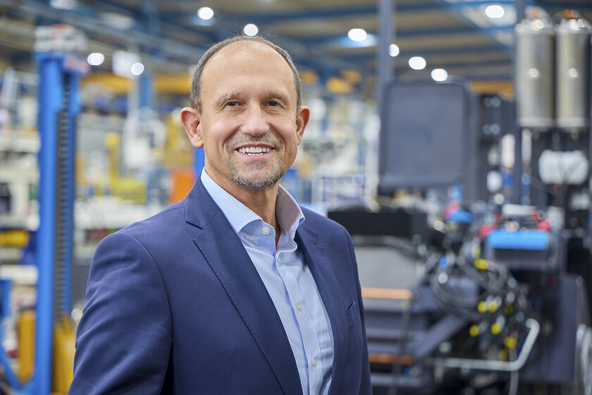 Ein freundlich lächelnder Mann mit einer fast glatze steht in einer Werkstatt. Er trägt einen blauen Anzug.