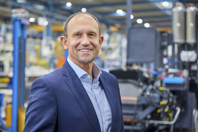 Ein freundlich lächelnder Mann mit einer fast glatze steht in einer Werkstatt. Er trägt einen blauen Anzug.