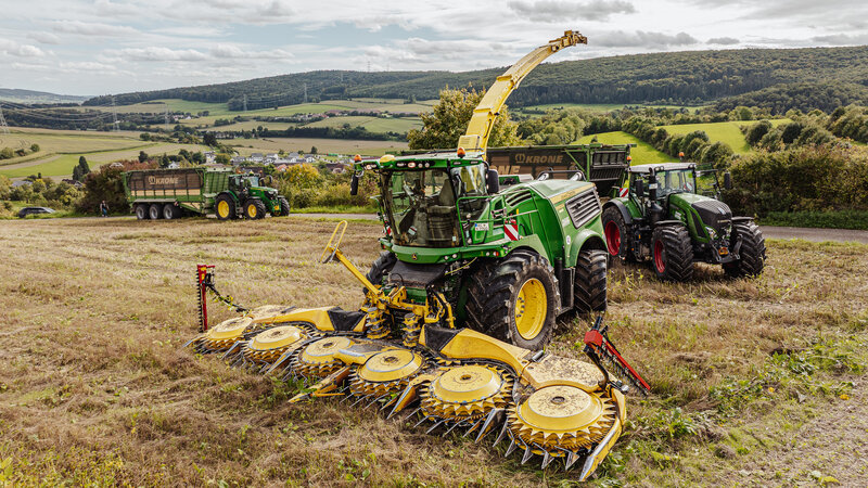 Drei Landwirtschaftliche Fahrzeuge stehen auf einem Feld. Vorne steht ein grün gelber Häcksler, dahinter stehen zwei weitere Traktoren jeweils mit einem großen Anhänger.