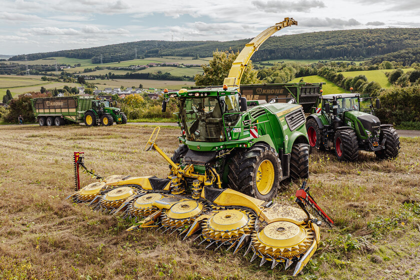 Drei Landwirtschaftliche Fahrzeuge stehen auf einem Feld. Vorne steht ein grün gelber Häcksler, dahinter stehen zwei weitere Traktoren jeweils mit einem großen Anhänger.