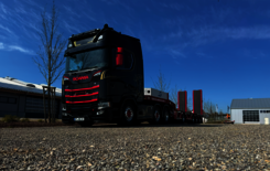 Ein schwarz roter Scania S 770 mit rot graunen Tieflader steht auf einem Schotterparkplatz neben einer Halle bei strahlend blauem Himmel.