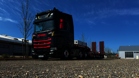 Ein schwarz roter Scania S 770 mit rot graunen Tieflader steht auf einem Schotterparkplatz neben einer Halle bei strahlend blauem Himmel.