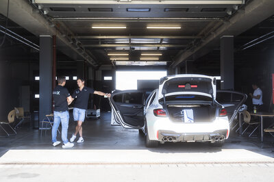 In einer dunklen mit Sonnenschein beleuchteten Garage stehen links zwei Männer die sich miteinander unterhalten. Einer der beiden hebt sich mit seiner linken Hand an einer offenen Tür eines weißen Autos fest, wo der Kofferraum offen ist und man ein NX NextMotion Zentralsteuergerät verbaut sieht.