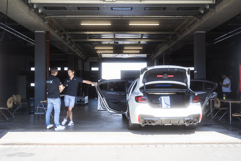 In einer dunklen mit Sonnenschein beleuchteten Garage stehen links zwei Männer die sich miteinander unterhalten. Einer der beiden hebt sich mit seiner linken Hand an einer offenen Tür eines weißen Autos fest, wo der Kofferraum offen ist und man ein NX NextMotion Zentralsteuergerät verbaut sieht.