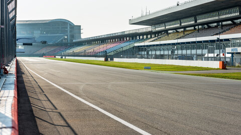 Die Start- und Zielgerade des Hockenheimrings bei Sonnenschein. Von Richtung Zaun schaut man zu den Zuschauertribünen, wo kein Mensch zu sehen ist, auch auf der Rennstrecke ist nichts los.