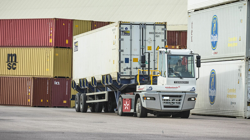 Ein Terminal Traktor von Terberg fährt mit einem Container auf dem Anhänger auf einem Gelände, wo noch weiter Container stehen.