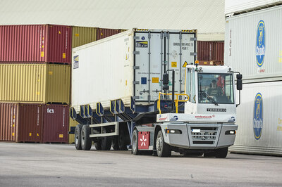 Ein Terminal Traktor von Terberg fährt mit einem Container auf dem Anhänger auf einem Gelände, wo noch weiter Container stehen.