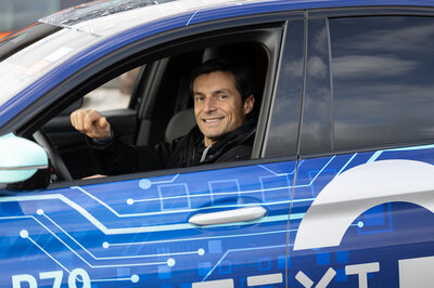 In einem gebrandeten blauen Auto ist bei der Fahrertür das Fenster unten. Im Auto sitzt ein lächelnder braunhaariger Mann und schaut aus dem Fenster. 
