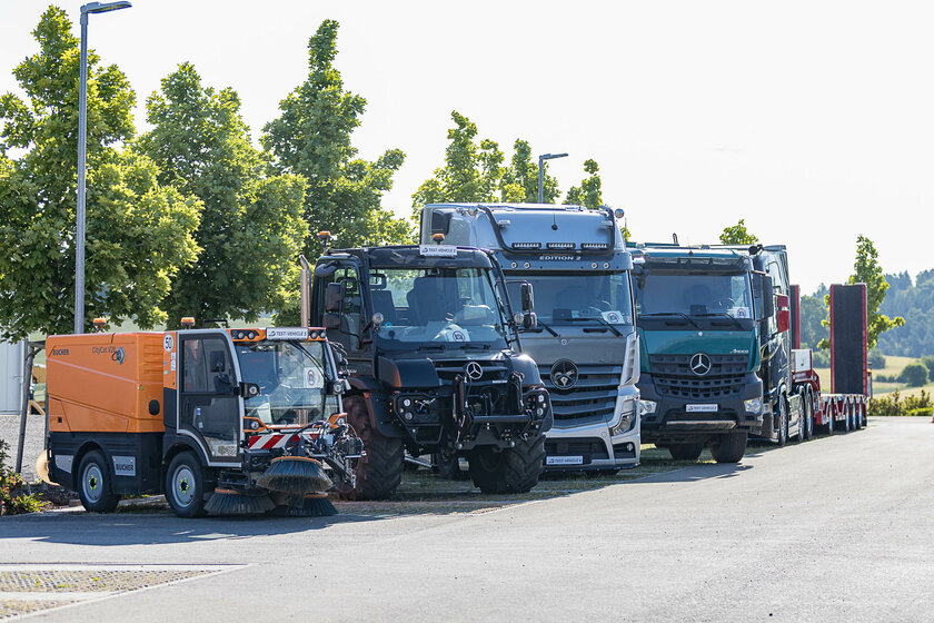 Reihe verschiedener Nutzfahrzeuge, darunter Straßenkehrmaschine, Unimog und mehrere Lkw von Mercedes-Benz, nebeneinander geparkt vor einer Baumreihe.