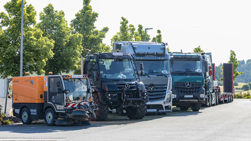 Reihe verschiedener Nutzfahrzeuge, darunter Straßenkehrmaschine, Unimog und mehrere Lkw von Mercedes-Benz, nebeneinander geparkt vor einer Baumreihe.