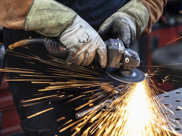 Zwei Hände mit Handschuhen halten eine Flex und flexen eine Metallstange ab. Es gibt sehr viele orange/gelbe funken.