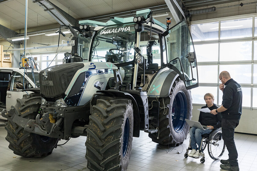 Ein Traktor steht in einer Halle. Rechts neben dem Traktor stehen zwei Menschen, Welche gerade etwas besprechen. Einer der zwei Männer sitzt im Rollstuhl.