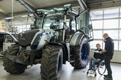 Ein Traktor steht in einer Halle. Rechts neben dem Traktor stehen zwei Menschen, Welche gerade etwas besprechen. Einer der zwei Männer sitzt im Rollstuhl.