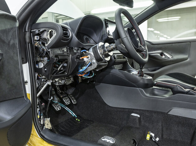 Eine Nahaufnahme, aus Sicht der Fahrertür, vom Lenkrad bis zum Gaspedal, indem ein Motor hinter dem Lenkrad neu verbaut ist, wo normal die Lenksäule verläuft.