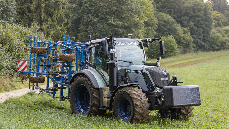 Ein silberblauer Traktor mit einem blauen Anbaugerät steht auf einer Wiese. Die Reifen und das Anbaugerät sind mit Erde bedeckt. 